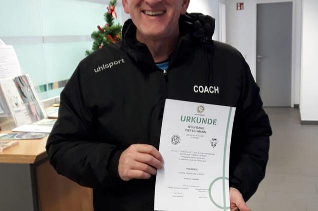 Mann in schwarzer Jacke hält stolz eine Trainerurkunde in der Hand, mit einem Lächeln im modernen Raum.