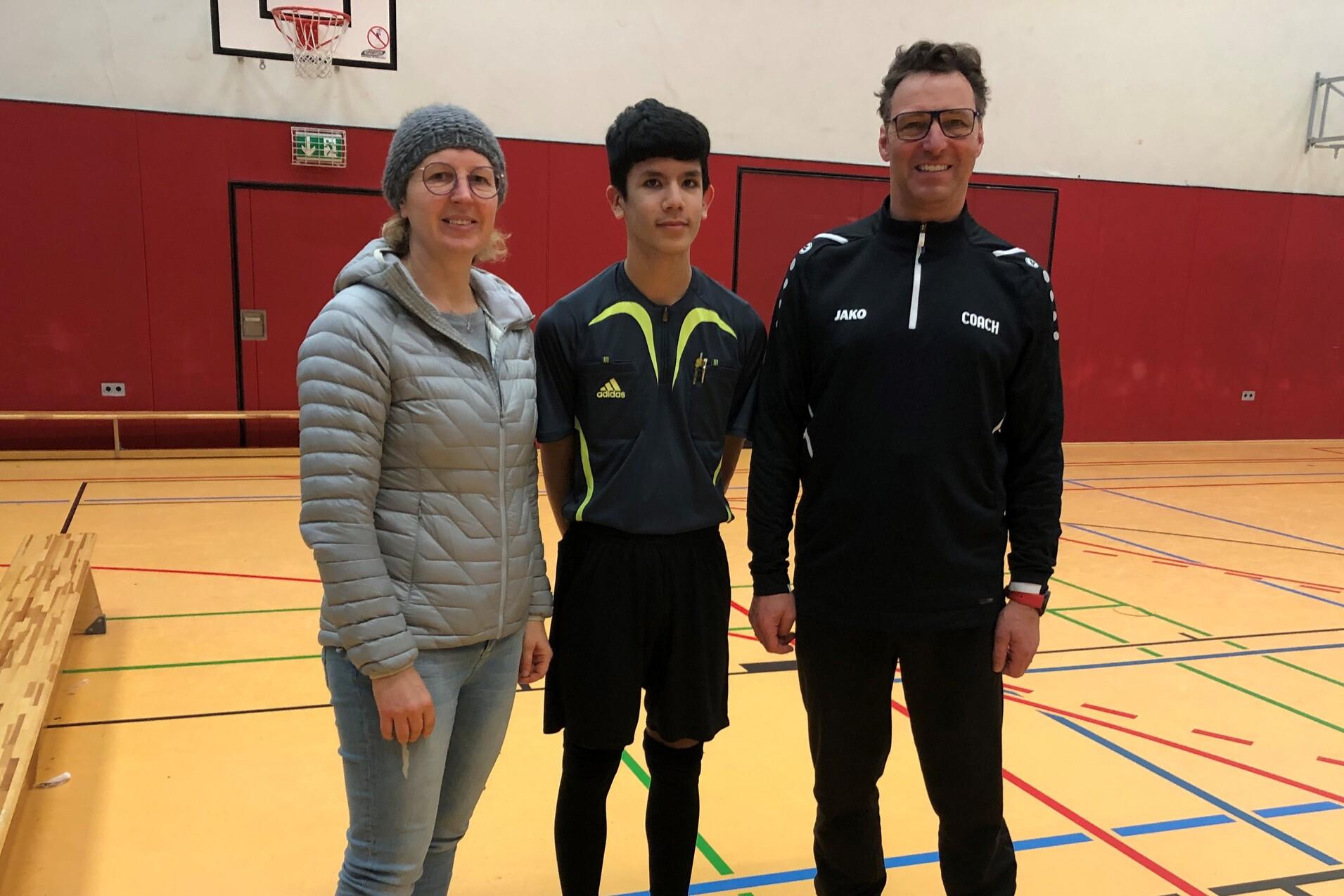 Drei Personen stehen in einer Sporthalle, eine Frau und zwei M&auml;nner in Sportkleidung, davor eine Bank und ein Basketballkorb.