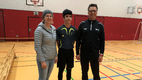 Drei Personen stehen in einer Sporthalle, ein Junge in Sportkleidung zwischen zwei Erwachsenen.