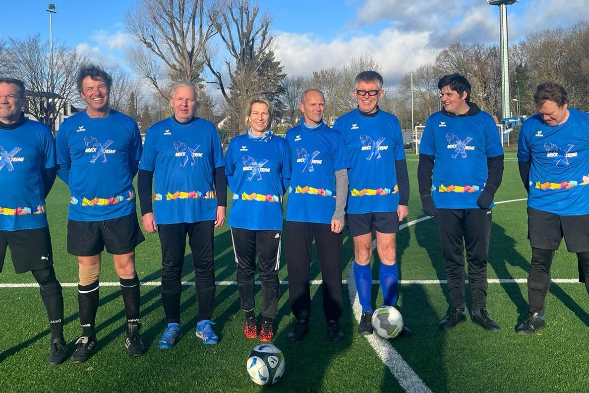 Gruppenfoto von sieben Fußballspielern in blauen Trikots auf einem grünen Platz mit Bäumen und blauem Himmel im Hintergrund.
