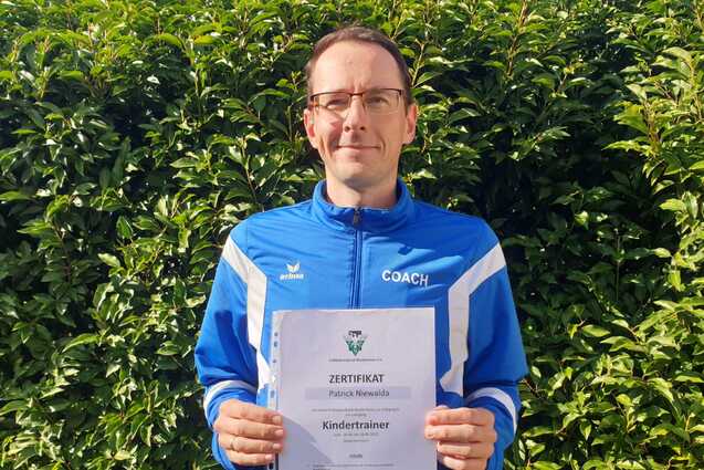 Mann h&auml;lt ein Zertifikat f&uuml;r die Trainerausbildung zum Kindertrainer vor einer gr&uuml;nen Hecke.