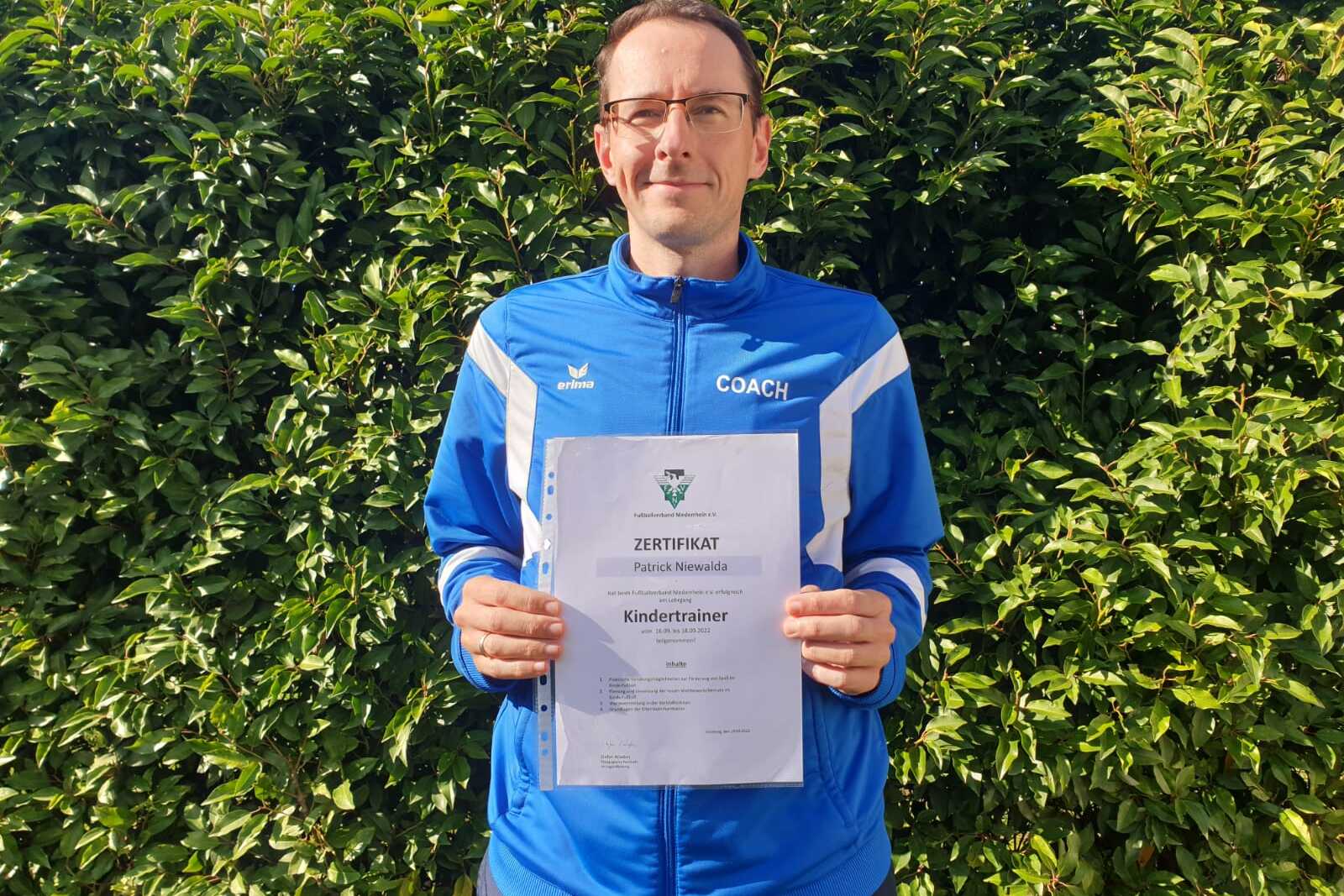 Mann hält ein Zertifikat für die Trainerausbildung zum Kindertrainer vor einer grünen Hecke.