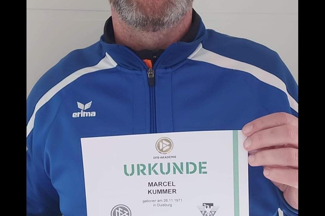 Mann mit Brille hält eine Trainer-C-Urkunde in den Händen, trägt einen blauen Trainingsanzug vor einem hellen Hintergrund.