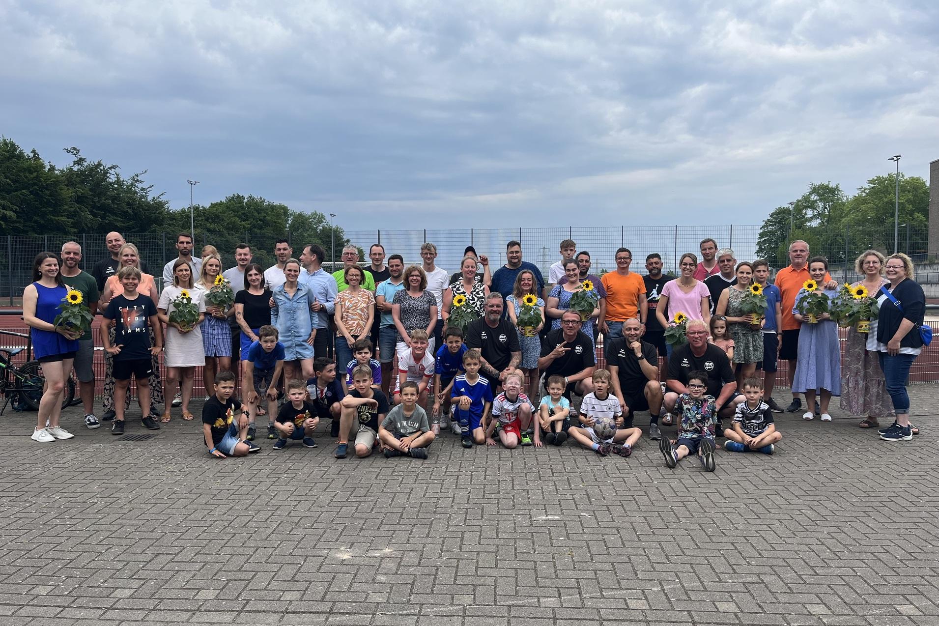 Gruppe von Personen unterschiedlichen Alters mit Sonnenblumen in der Hand, versammelt auf einem Platz nahe einem Tennisplatz.