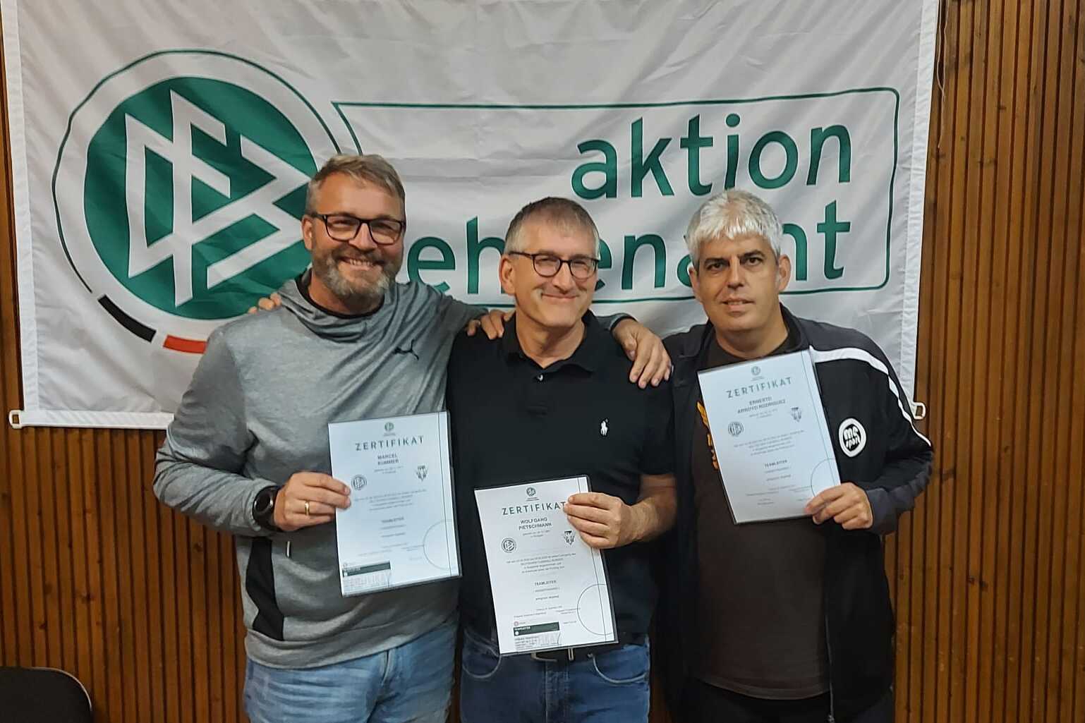 Drei Männer halten Urkunden in den Händen, stehend vor einem Banner mit der Aufschrift „aktion ehrenamt“.