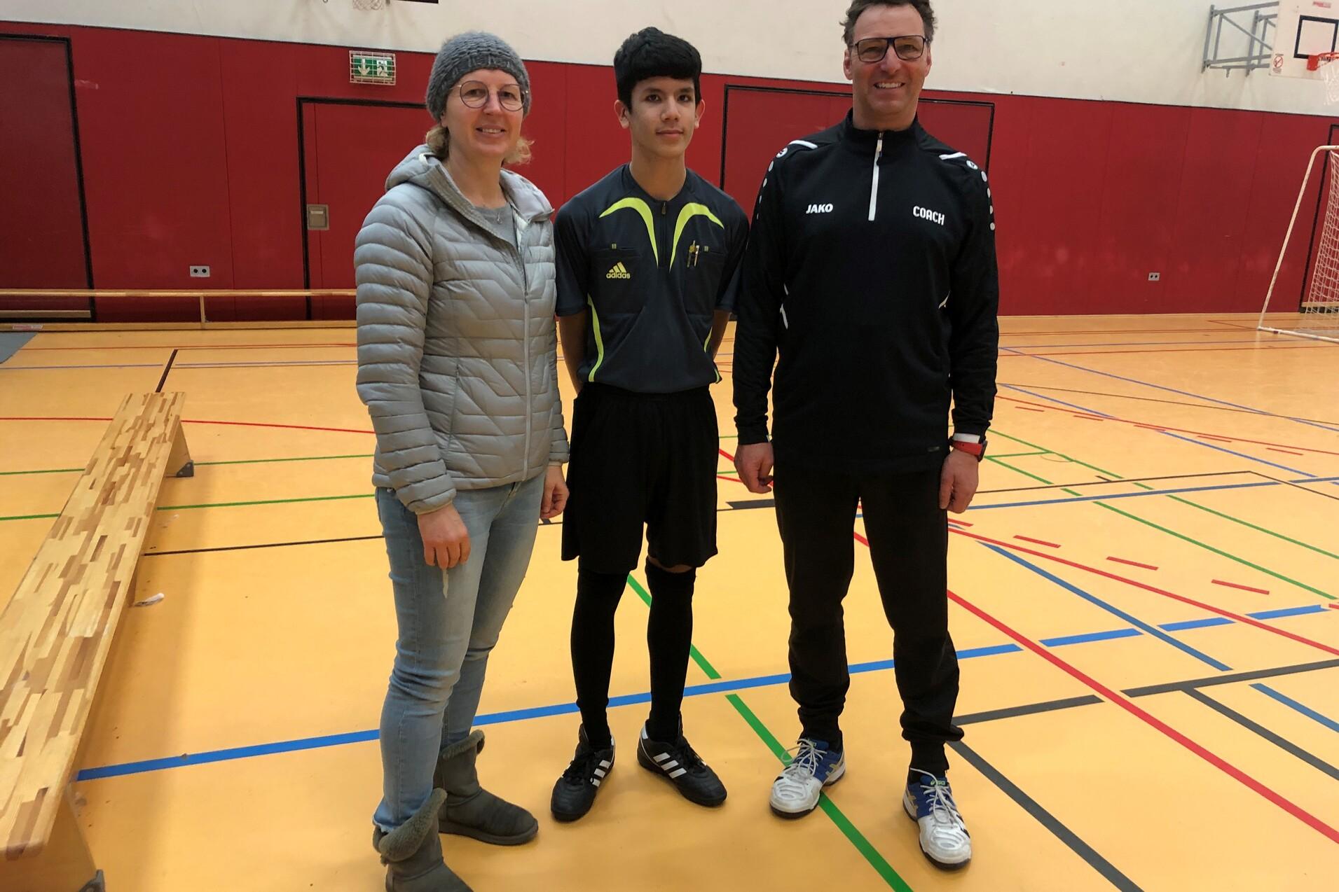 Drei Personen stehen in einer Sporthalle, eine Frau und zwei Männer in Sportkleidung, davor eine Bank und ein Basketballkorb.