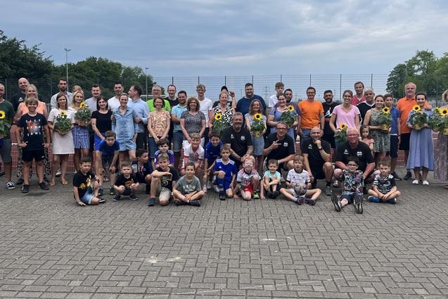 Gruppe von Personen unterschiedlichen Alters mit Sonnenblumen in der Hand, versammelt auf einem Platz nahe einem Tennisplatz.