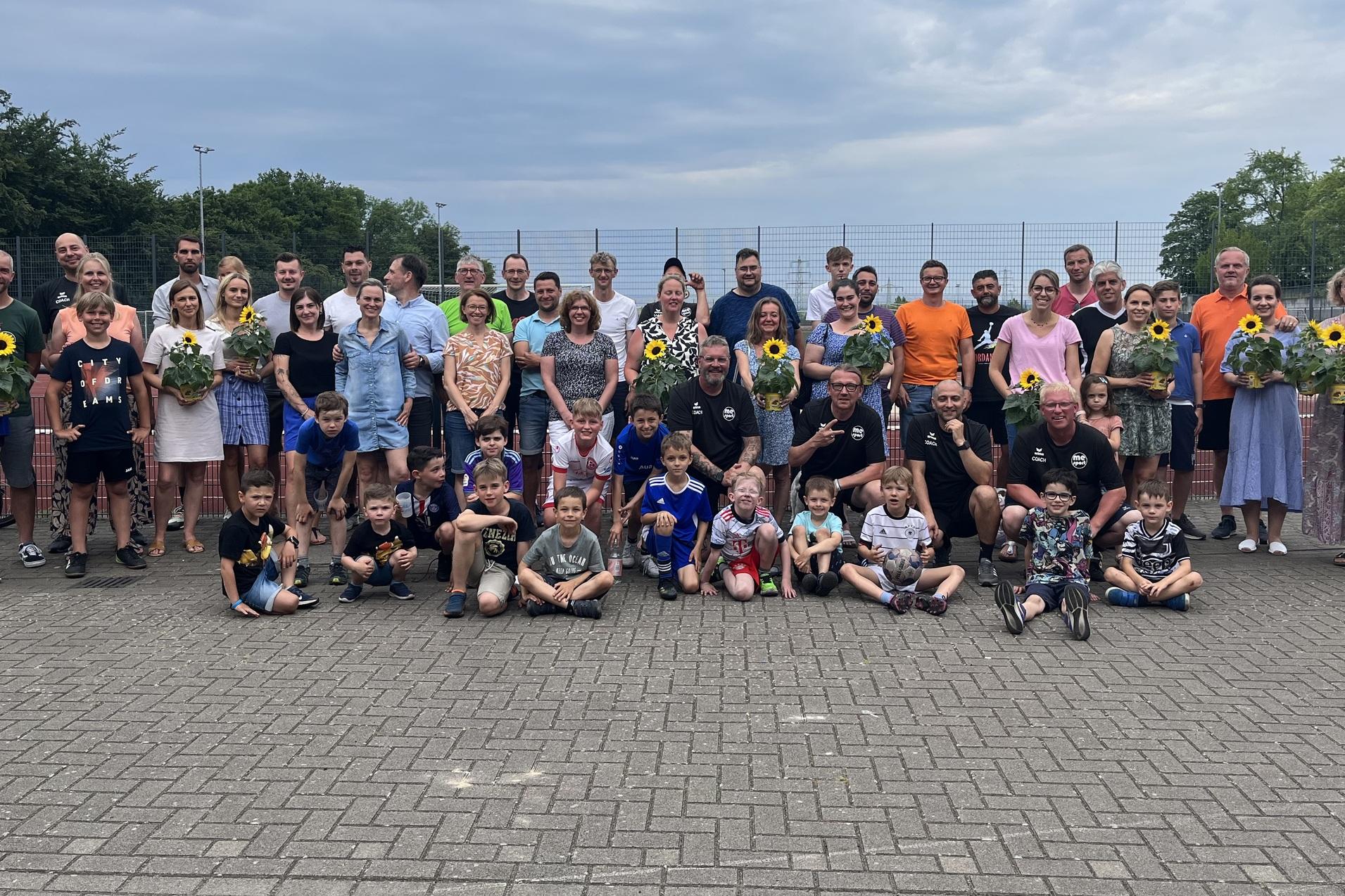 Gruppe von Personen unterschiedlichen Alters mit Sonnenblumen in der Hand, versammelt auf einem Platz nahe einem Tennisplatz.