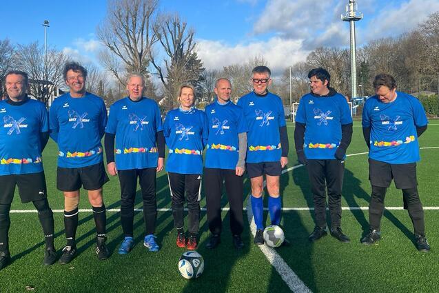 Gruppenfoto von sieben Fu&szlig;ballspielern in blauen Trikots auf einem gr&uuml;nen Platz mit B&auml;umen und blauem Himmel im Hintergrund.