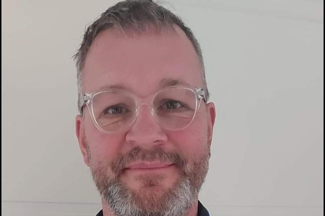 Mann mit Brille h&auml;lt eine Trainer-C-Urkunde in den H&auml;nden, tr&auml;gt einen blauen Trainingsanzug vor einem hellen Hintergrund.