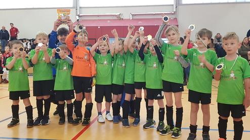 Kinderfußballmannschaft in grünen Trikots hält Medaillen und einen Snack nach einem Turnier stolz in die Höhe.