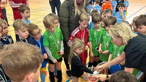 Eine Gruppe von Kindern steht um eine Erwachsene, die Preise in einer Sporthalle verteilt.
