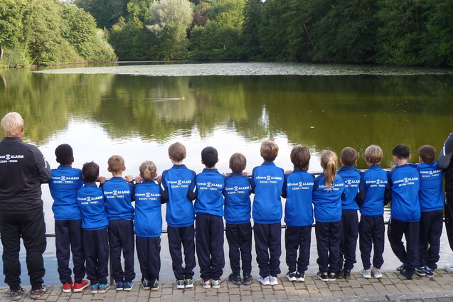 Gruppe von Kindern und Betreuern in blauen Sportjacken steht am Ufer eines ruhigen Teichs und blickt auf das Wasser.