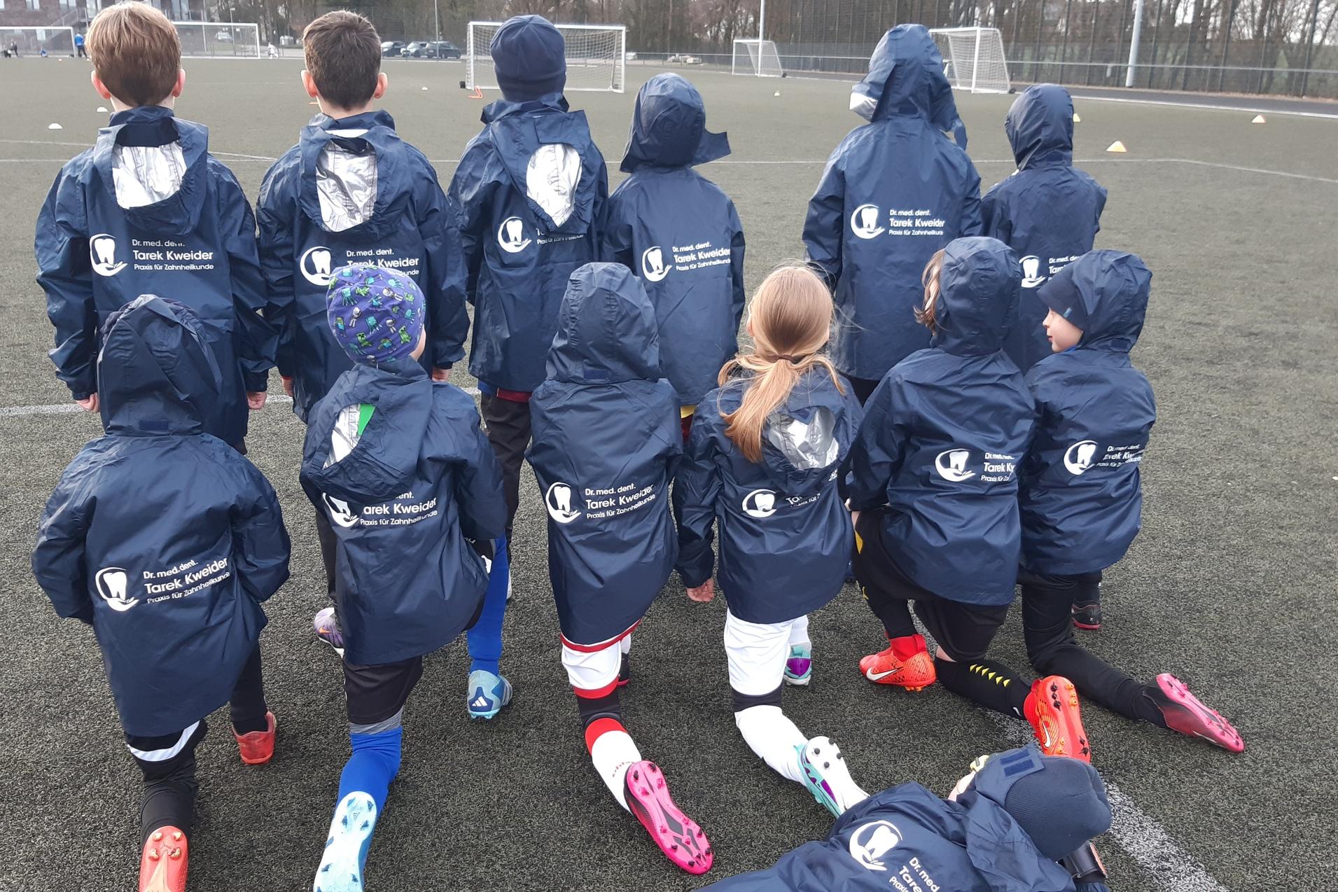 Gruppe von Kindern in blauen Trainingsjacken auf einem Fu&szlig;ballfeld, die von hinten auf das Spielfeld schauen.