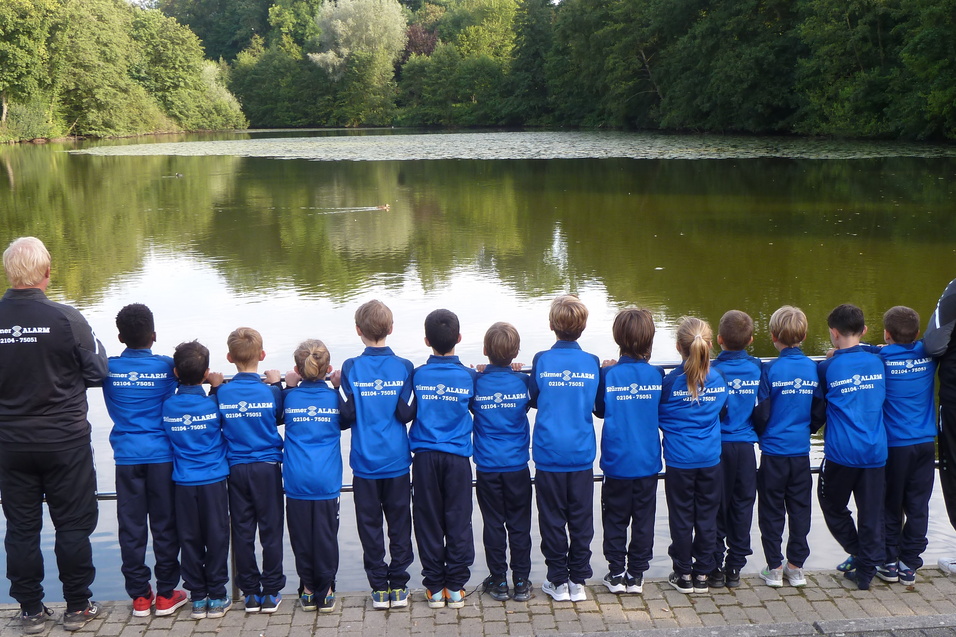 Gruppe von Kindern und Betreuern in blauen Sportjacken steht am Ufer eines ruhigen Teichs und blickt auf das Wasser.