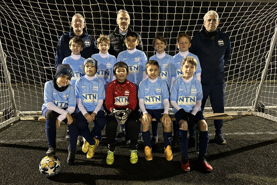Eine Fußballmannschaft aus Kindern in blauen Trikots steht vor einem Tor, zwei Trainer hinter ihnen.