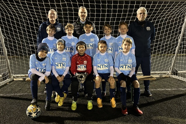 Eine Fußballmannschaft aus Kindern in blauen Trikots steht vor einem Tor, zwei Trainer hinter ihnen.