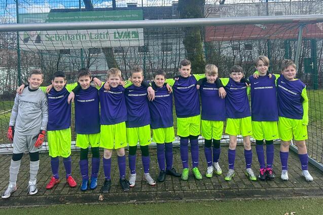 Gruppe von elf Jungen in Fußballtrikots, lächelnd vor einem Tor auf einem Sportplatz.