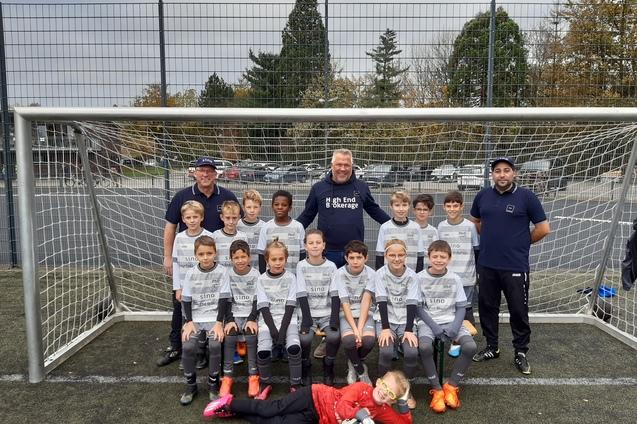 Gruppenfoto einer Fußballmannschaft von Kindern in Trikots, stehend vor einem Tor, mit drei Trainern im Hintergrund.