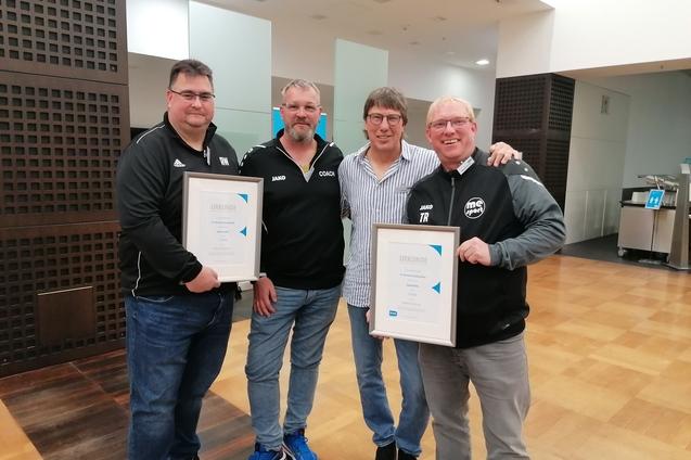 Vier Männer stehen in einem Innenraum und halten Urkunden in den Händen, lächelnd und in sportlicher Kleidung.