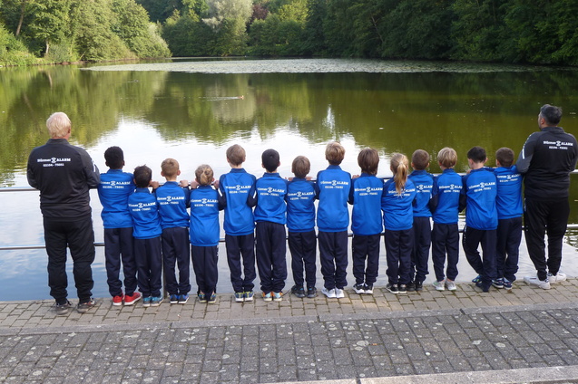 Gruppe von Kindern und Betreuern in blauen Sportjacken steht am Ufer eines ruhigen Teichs und blickt auf das Wasser.