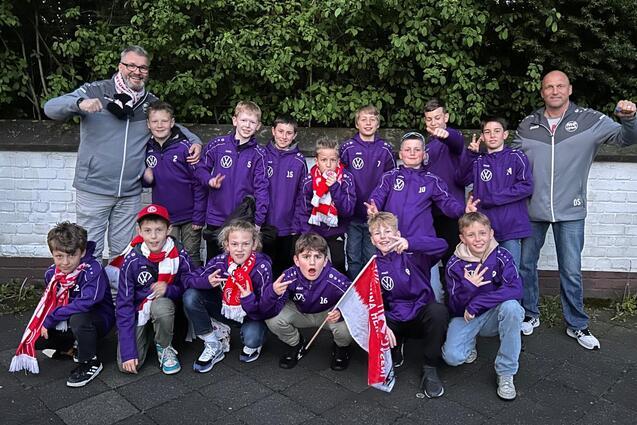 Gruppe von Kindern in lila Sportjacken und Schals posiert mit zwei Trainern im Freien vor einer grünen Hecke.