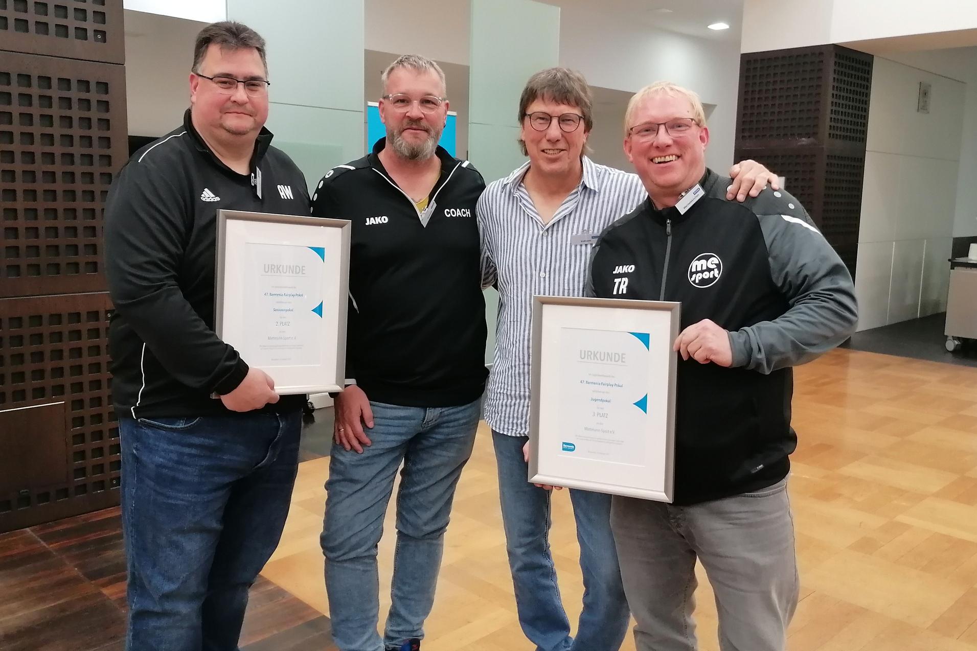 Vier M&auml;nner stehen in einem Innenraum und halten Urkunden in den H&auml;nden, l&auml;chelnd und in sportlicher Kleidung.