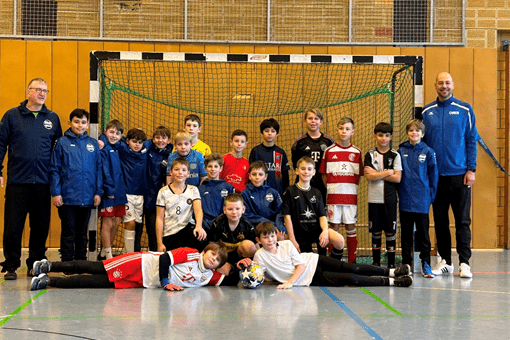 Gruppe von Kindern in Sportkleidung und zwei Trainern vor einem Fußballtor in einer Sporthalle.