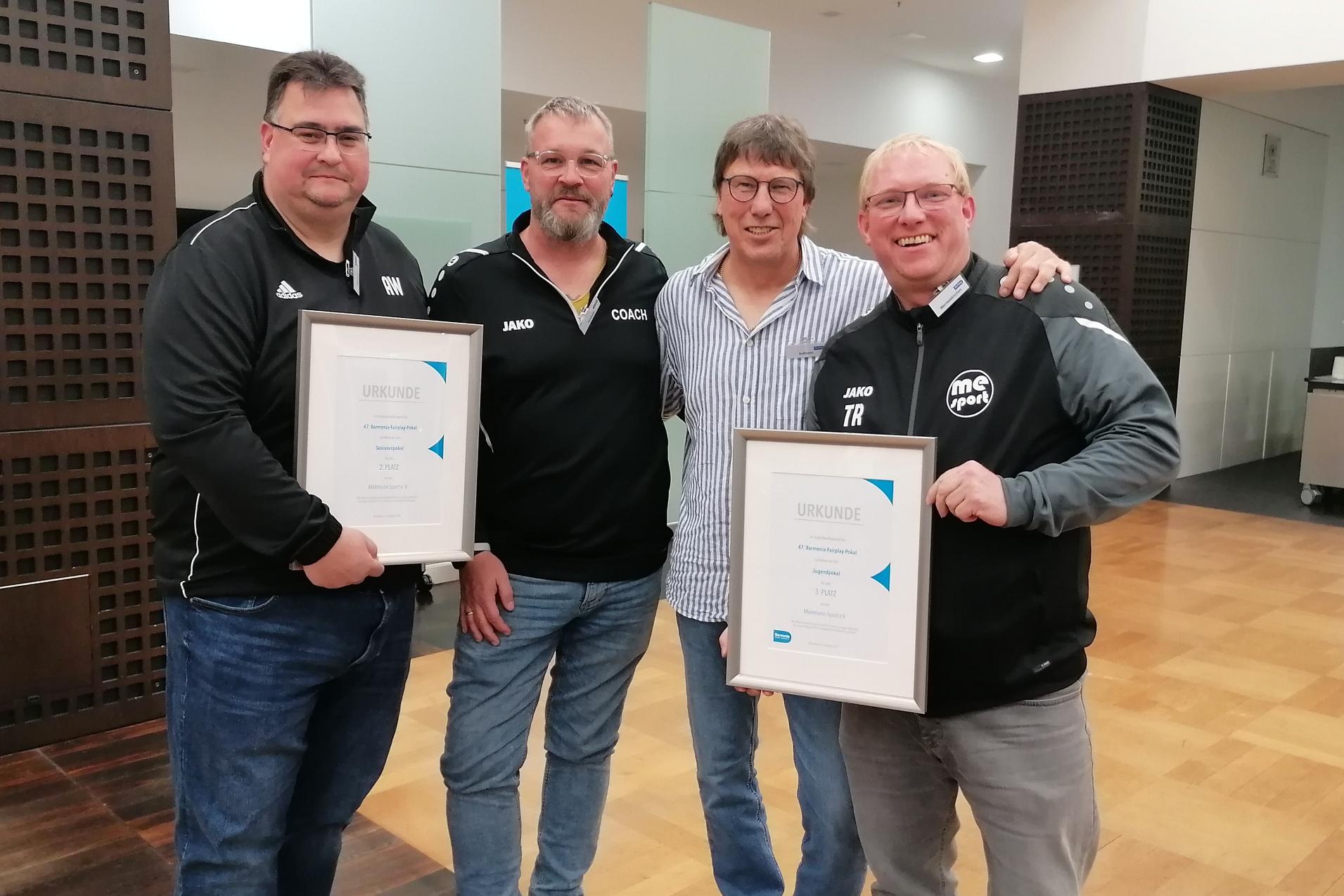 Vier M&auml;nner stehen in einem Innenraum und halten Urkunden in den H&auml;nden, l&auml;chelnd und in sportlicher Kleidung.