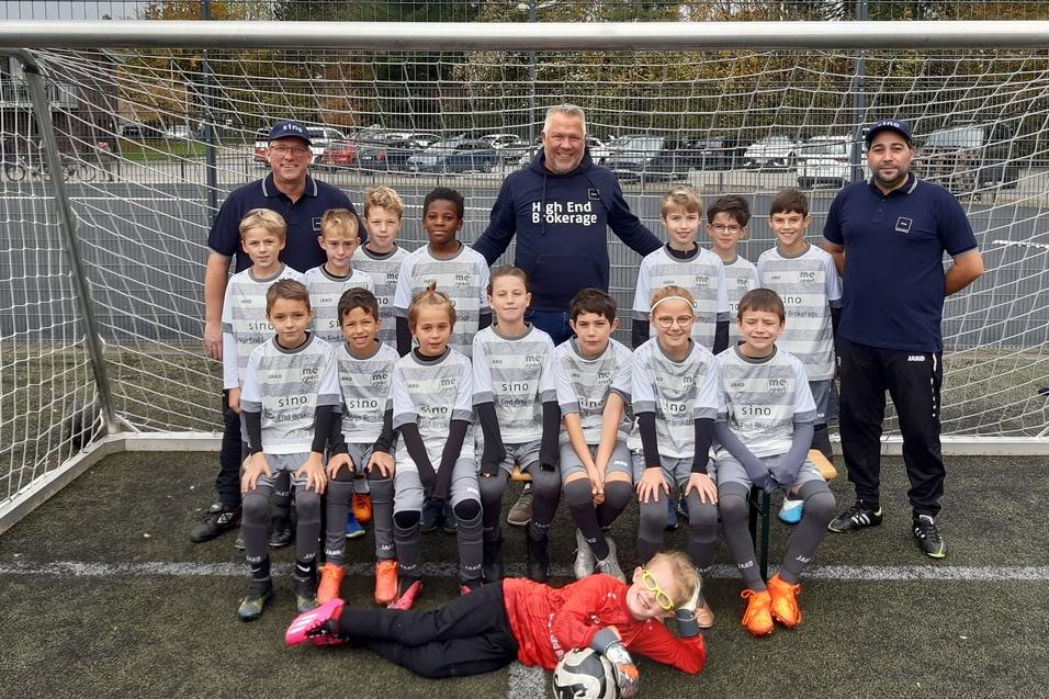 Gruppenfoto einer Fußballmannschaft von Kindern in Trikots, stehend vor einem Tor, mit drei Trainern im Hintergrund.