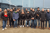 Gruppe von 16 Personen in winterlicher Kleidung vor einem modernen Gebäude, lächelnd und stehend auf dem Platz.