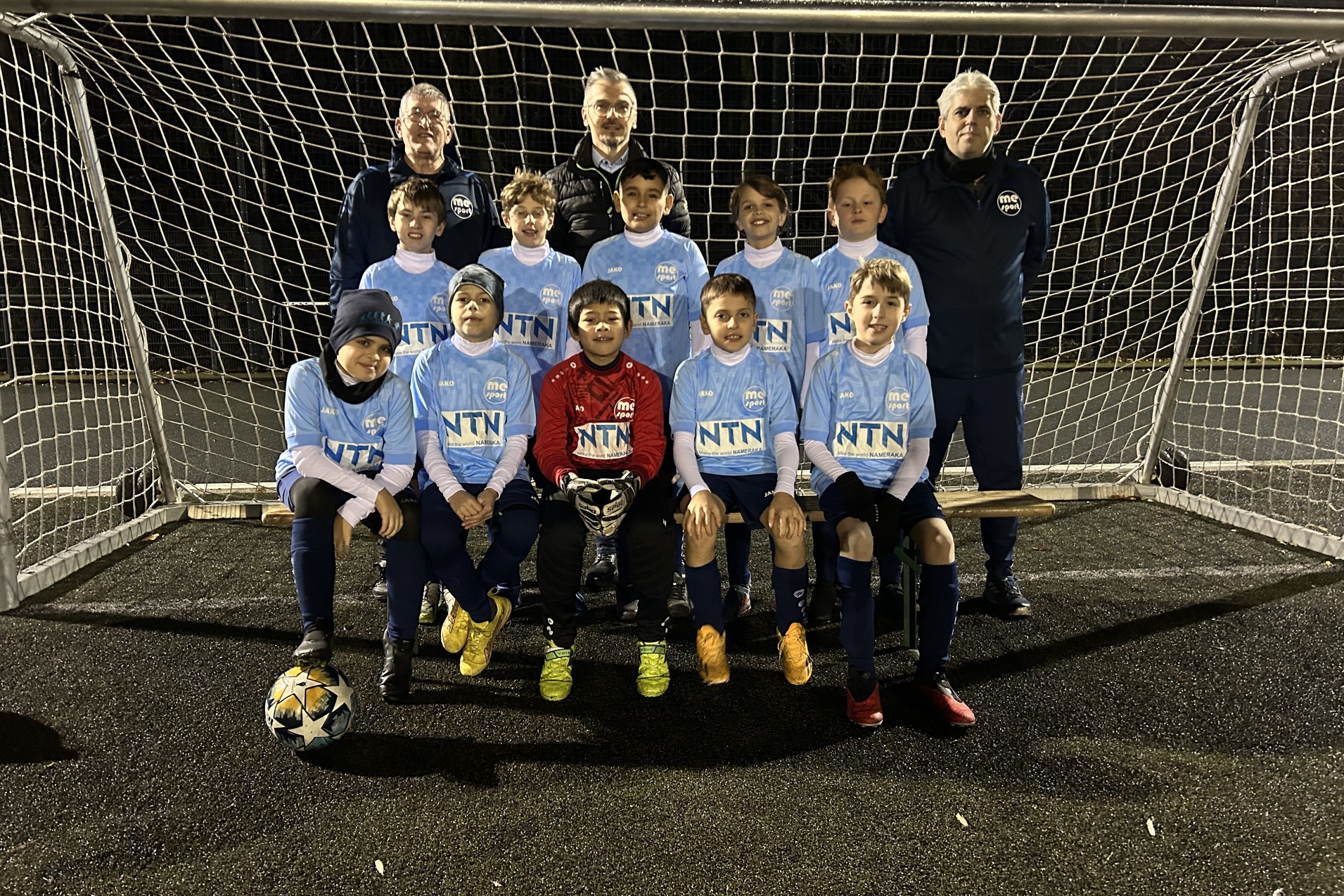Eine Fußballmannschaft aus Kindern in blauen Trikots steht vor einem Tor, zwei Trainer hinter ihnen.