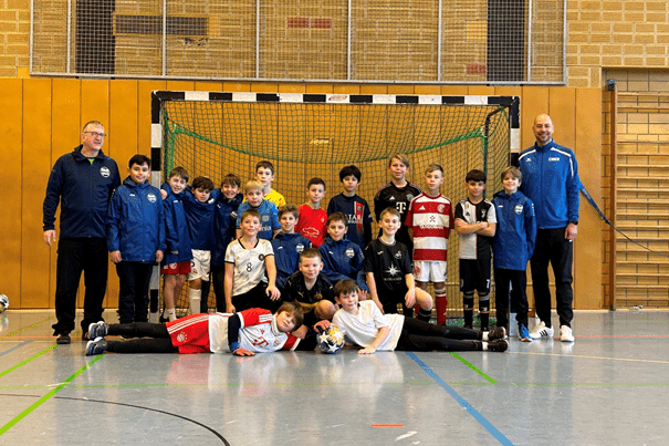 Gruppe von Kindern in Sportkleidung und zwei Trainern vor einem Fußballtor in einer Sporthalle.