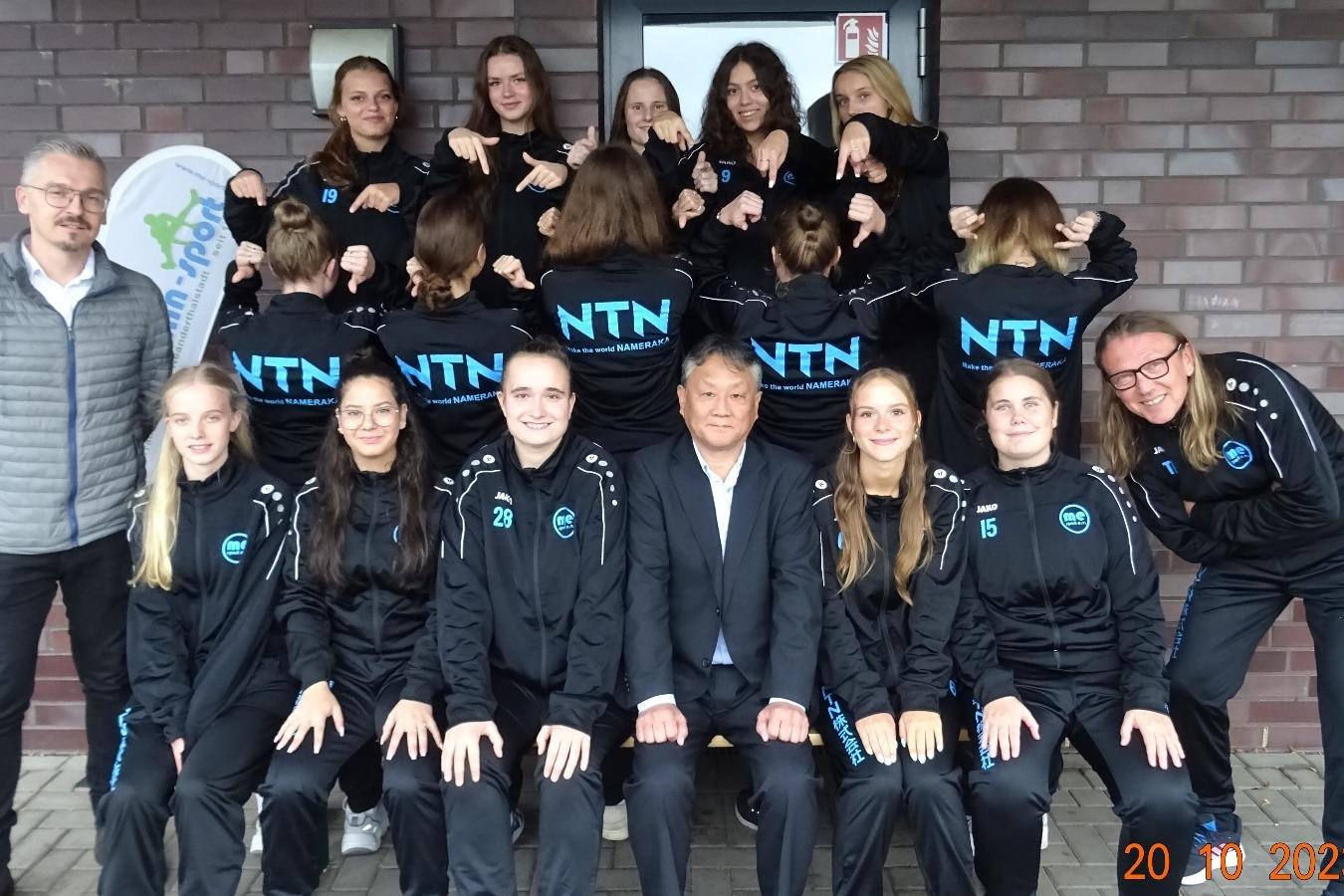 Gruppenfoto einer Mannschaft in schwarzen Trainingsanzügen mit „NTN“-Aufdruck vor einer Backsteinmauer.