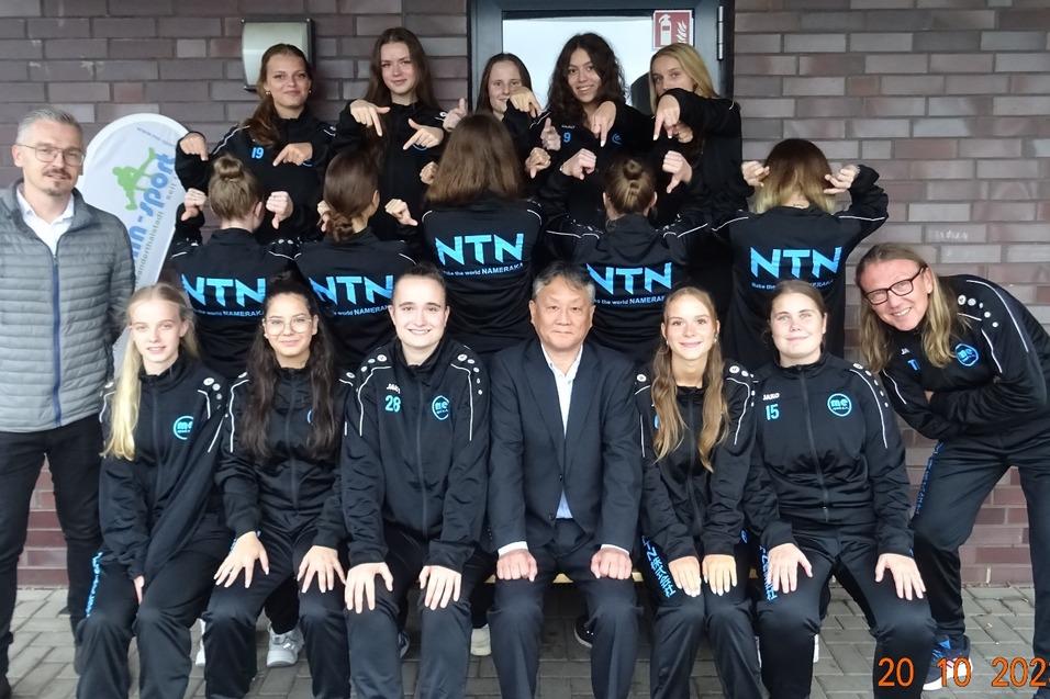 Gruppenfoto einer Mannschaft in schwarzen Trainingsanzügen mit „NTN“-Aufdruck vor einer Backsteinmauer.