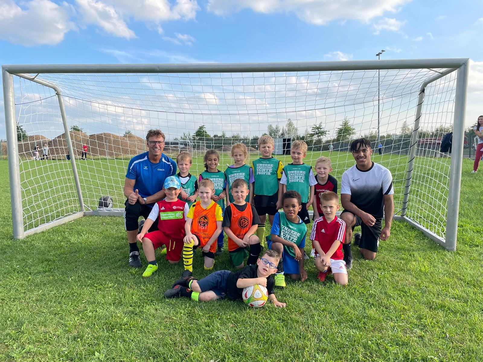 Gruppe von Kindern in Fußballtrikots posiert lächelnd vor einem Tor auf einem Rasenplatz, trainiert von zwei Erwachsenen.