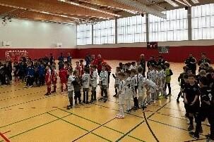 Versammlung von Kinderfußballmannschaften in Sportkleidung in einer Halle mit großen Fenstern und einem Fußballtor.