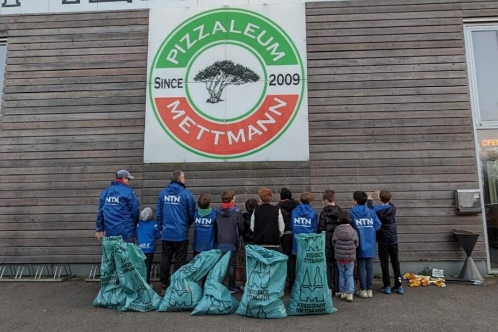 Gruppe von Kindern und Erwachsenen in blauen Jacken stehen vor einem Pizzarestaurant mit einer gro&szlig;en Werbetafel.