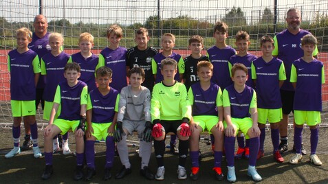 Mannschaftsfoto einer Jugendfußballmannschaft in lila-neonfarbenen Trikots, stehend und sitzend vor einem Tor.