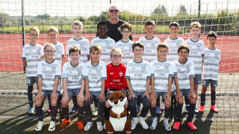 Mannschaftsfoto einer Jugendfußballmannschaft mit Trainer vor einem Tor, einige Kinder halten ein Maskottchen.