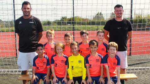 Kinderfußballmannschaft posiert mit Trainern vor einem Fußballtor auf einem Kunstrasenplatz.