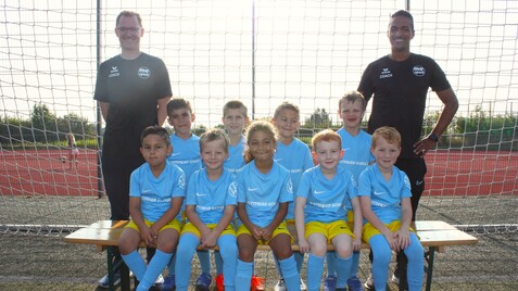 Jugendfußballmannschaft mit Trainer, alle tragen hellblaue Trikots und stehen vor einem Fußballtor.