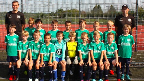 Gruppenfoto einer Jugendfußballmannschaft mit Trainer, alle tragen grüne Trikots und stehen vor einem Fußballtor.