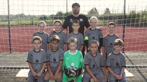 Fußballmannschaft von Kindern in grauen Trikots und Trainer, auf dem Sportplatz vor einem Tor, lächelnd posierend.