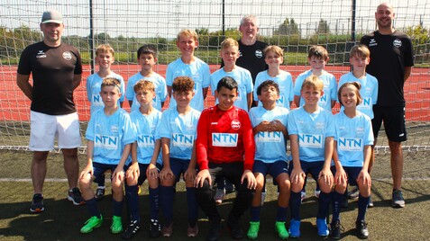 Gruppe von jungen Fußballspielern in blauen Trikots posiert vor einem Tor auf einem Sportplatz, zwei Trainer stehen hinten.