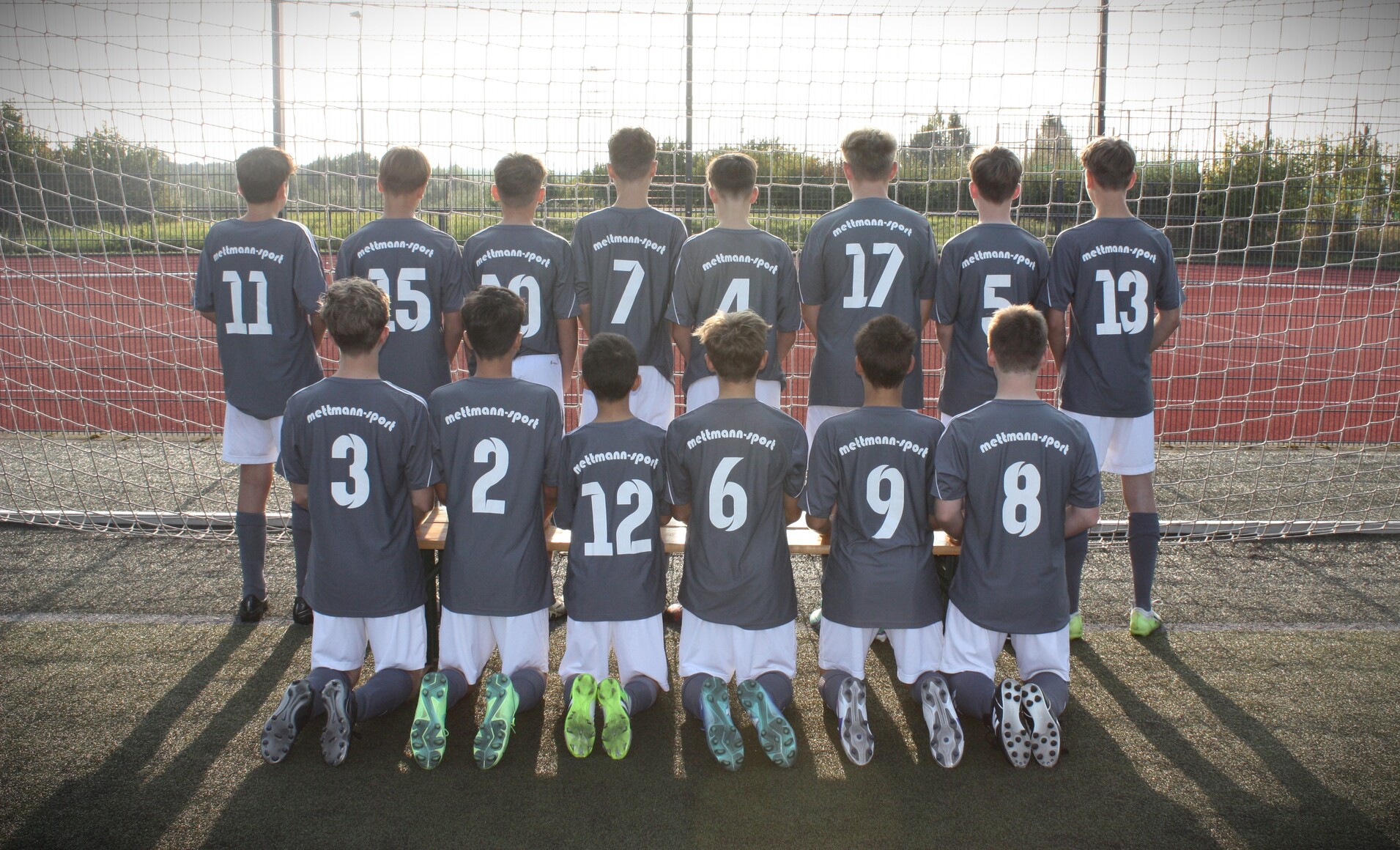 Junge Fußballmannschaft kniet hinter einer Bank, trägt graue Trikots mit Nummern und dem Schriftzug „meltman-sport“.