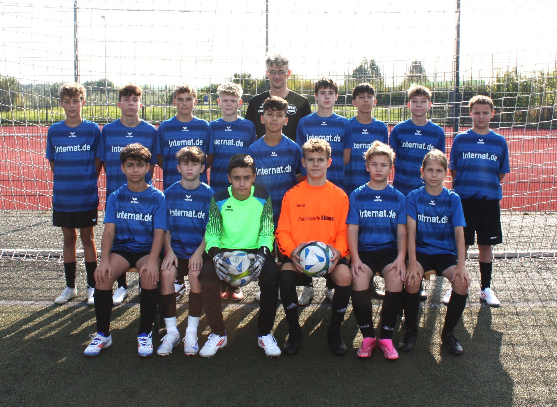 Ein Jugendfußballteam in blauen Trikots steht vor einem Tor auf einem Sportplatz, drei Spieler halten Bälle.