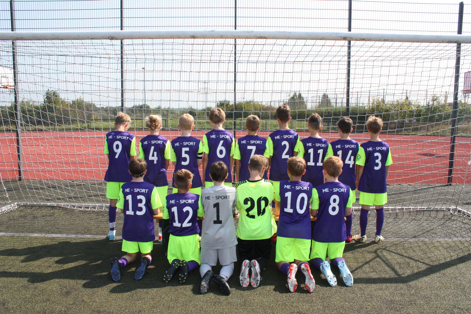 Gruppe von elf Fußballspielern und einem Torwart in lila-grünen Trikots vor einem Fußballtor auf einem Kunstrasenplatz.
