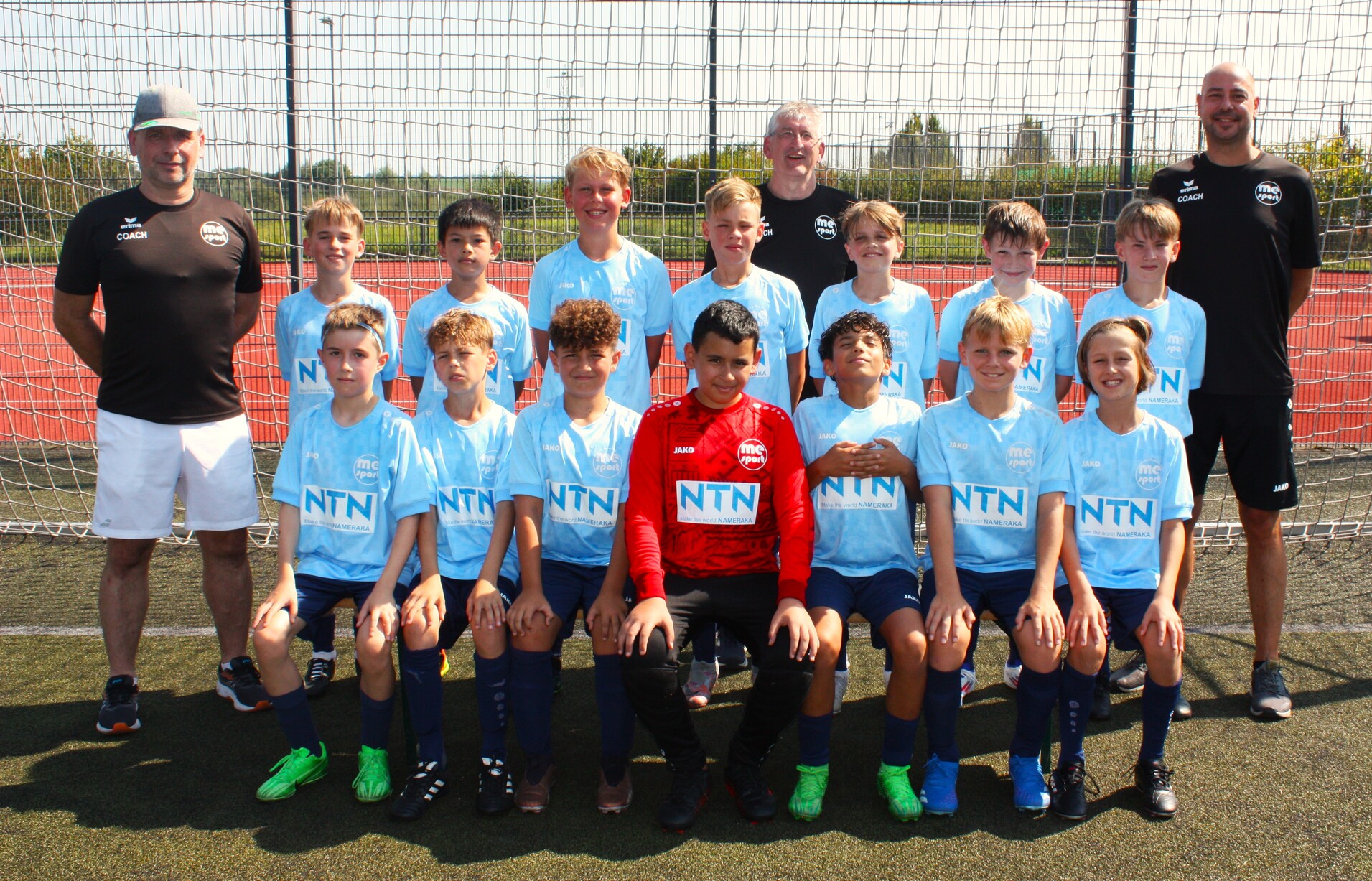 Gruppe von jungen Fußballspielern in blauen Trikots posiert vor einem Tor auf einem Sportplatz, zwei Trainer stehen hinten.