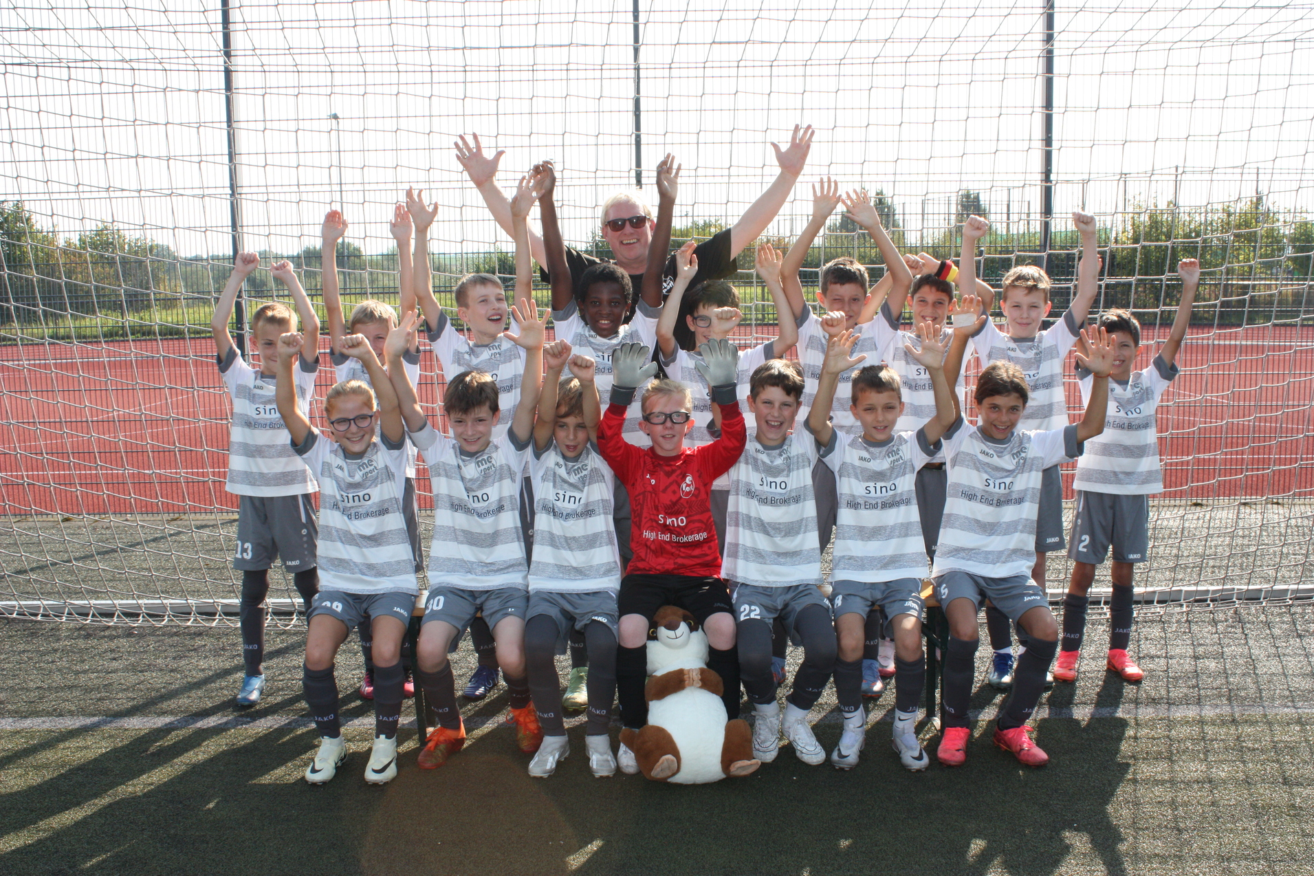 Gruppenselfie von Kindern in Fußballtrikots, die vor einem Tor stehen, und ein Kind hält einen Stofftierhund.